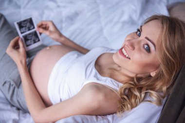 Sonogram holding, kameraya bakıyor ve yatakta yatarken gülümseyen güzel hamile kadının yüksek açılı görünüş