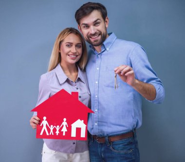 Mutlu güzel çift holding kağıt ev ve kameraya bakıyor ve gülümsüyor, gri arka plan üzerinde tuşları,