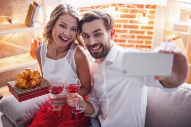 Güzel genç çift bardak şarap holding, selfie yapıyor ve süre evde kutlama gülümseyen