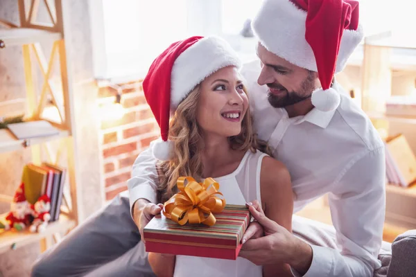美しい若い女性とサンタの帽子の男は家でクリスマスを祝っています 男は彼のガール フレンドにギフト ボックスを与えています — ストック写真