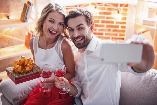 Beautiful young couple is holding glasses of wine, doing selfie and smiling while celebrating at home