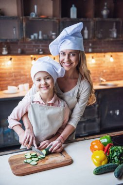 Sevimli küçük kız ve Şef'in şapka onun güzel annem sebze kesme ve evde mutfakta yemek yaparken gülümseyen