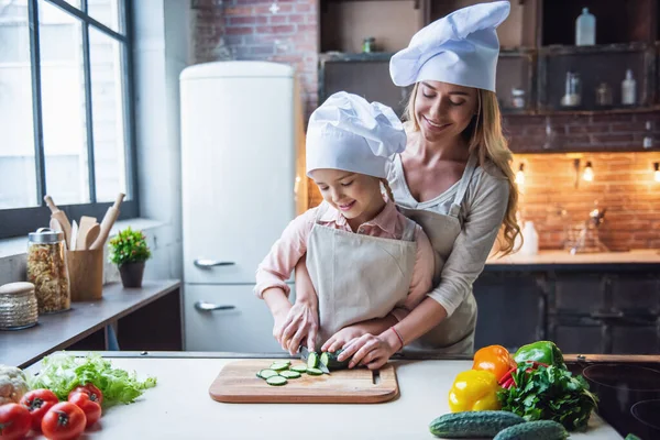Cute little girl and her beautiful mom in chef\'s hats are cutting vegetables and smiling while cooking in kitchen at home