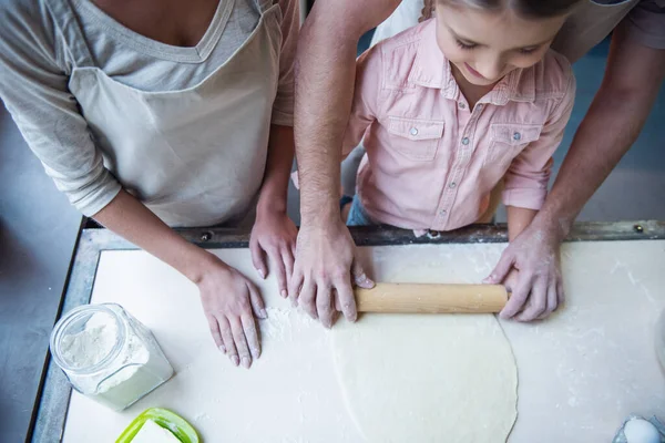 Top view of cute little girl and her beautiful parents flattening dough while baking in kitchen at home