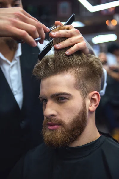 Handsome bearded man is looking forward while getting haircut by hairdresser at the barbershop