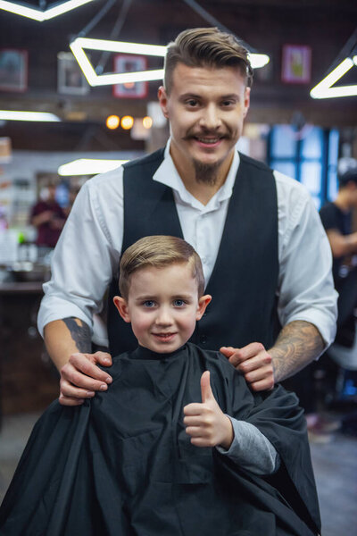Cute little boy is showing Ok sign and smiling after having his haircut done by hairdresser at the barbershop