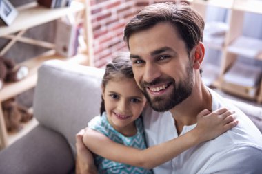Sevimli küçük kız ve yakışıklı babası sarılma, kameraya bakıyor ve evde kanepede otururken gülümseyen