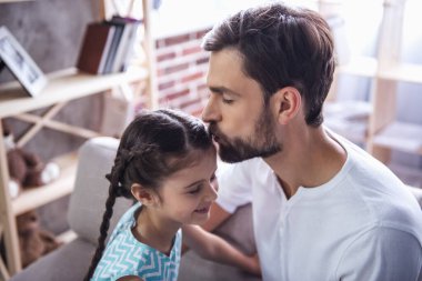 Yakışıklı genç Baba sevimli küçük kızını Alnından öpüyor. Her ikisi de evde kanepede oturuyor