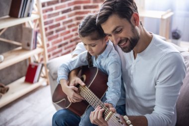 Sevimli küçük kız ve yakışıklı babası gitar çalmak ve evde kanepede otururken gülümseyen