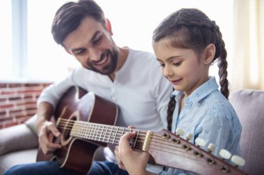 Sevimli küçük kız ve yakışıklı babası gitar çalmak ve evde kanepede otururken gülümseyen