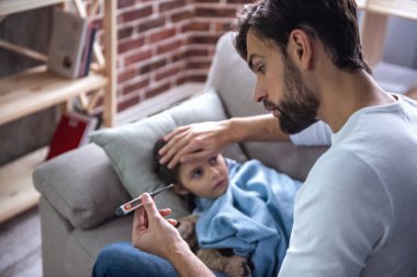 Babası ısısı alıyor iken battaniyeye örtülü hasta küçük kız kanepede yatıyor