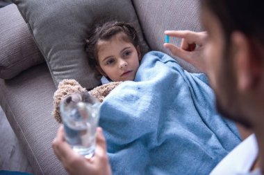 Babası haplarını ve bir bardak su veriyor iken battaniyeye örtülü hasta küçük kız kanepede yatıyor