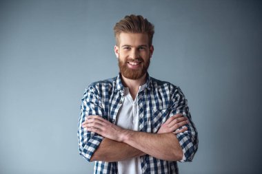 Rahat kıyafetler içinde yakışıklı sakallı adam kameraya bakıyor ve gri arka plan üzerinde katlanmış silah ile dururken gülümseyen