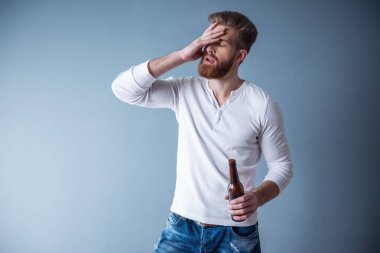 Yakışıklı sakallı adam rahat kıyafetler içinde bir şişe içecek ve gri arka plan üzerinde gözlerini kapsayan tutuyor