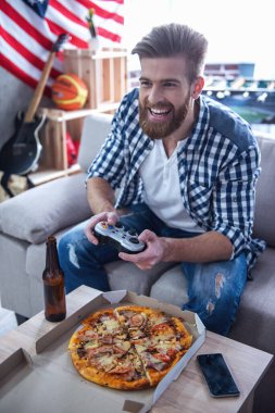 Sakallı yakışıklı olan video oyunları oynarken, bira içiyor, pizza yemek yiyor ve evde kanepede otururken gülümseyen