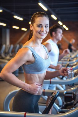 Çekici genç kadın kameraya bakarak ve spor salonunda bir koşu bandı üzerinde çalıştırırken gülümseyen
