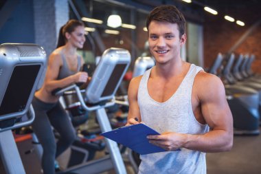 Yakışıklı kaslı eğitmen kameraya bakıyor ve çekici genç kadın üzerinde eliptik bir eğitmen spor salonunda egzersiz arka planda gülümseyen