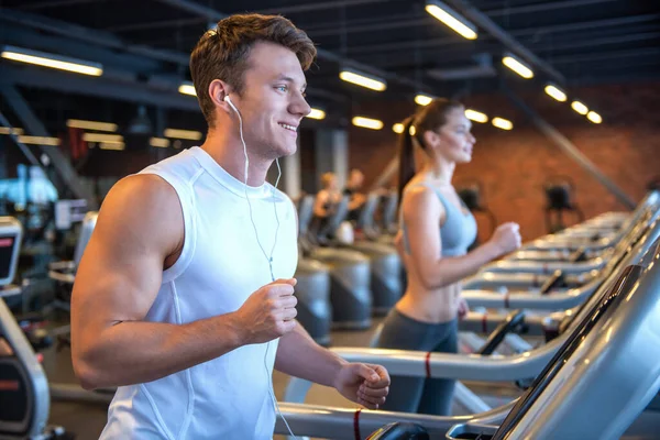 Attractive young muscular man in earphones is smiling while running on a treadmill in gym