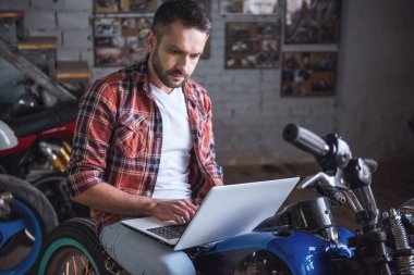 Yakışıklı adam rahat kıyafetler içinde motosiklet tamir dükkanında otururken bir laptop kullanıyor