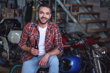 Yakışıklı adam rahat kıyafetler içinde bir şişe bira, kamera ve motosiklet tamir dükkanında otururken tutuyor