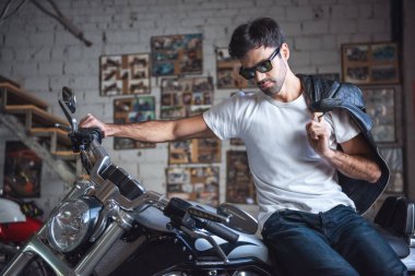 Güneş gözlüklü çekici kendine güvenen adam onun deri ceket tutarak ve motosiklet tamir dükkanında otururken bakmak