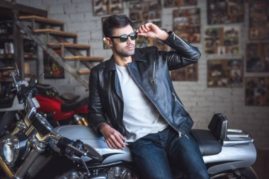 Deri ceket ve güneş gözlüklü çekici kendine güvenen adam uzakta üstünde motosiklet tamir dükkanında otururken arıyor