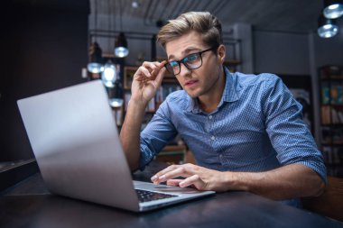 Gözlük çekici genç adam modern kütüphanede otururken bir laptop kullanıyor