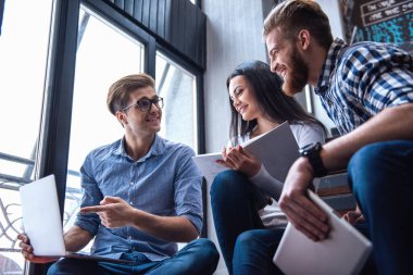 Genç akıllı insanlar becerikli alet ve kitaplar, söz ve modern kütüphanede merdivenlerde otururken gülümseyen kullanıyorsanız