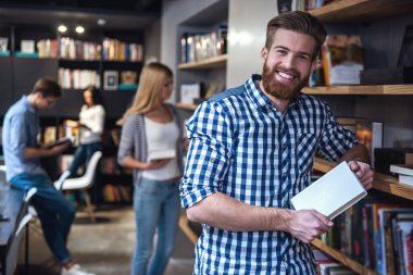 Genç akıllı insanlar modern kütüphanede çalışıyoruz. Adam ön planda kameraya bakarak ve gülümseyen bir kitap tutuyor