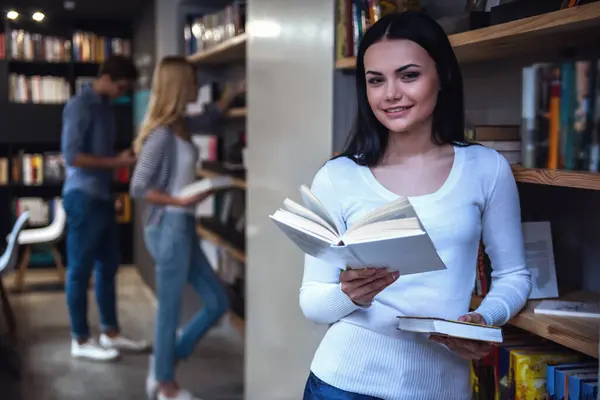 Genç akıllı insanlar modern kütüphanede çalışıyoruz. Kameraya bakarak ve gülümseyen kız ön planda bir kitap tutuyor