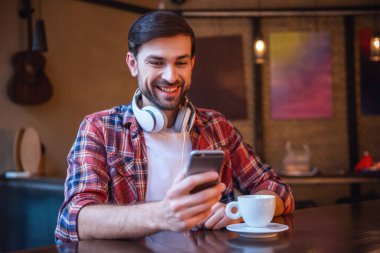 Yakışıklı adam kulaklık ile bir smartphone kullanarak, kahve içme ve kafede istirahat ederken gülümseyen