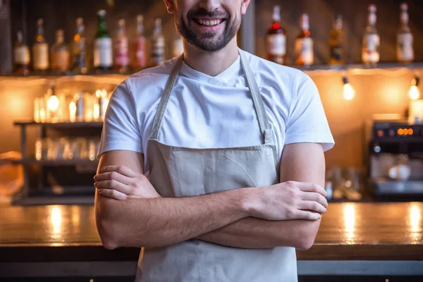 Cropped image of handsome barista in apron smiling while standing with crossed arms near the bar counter in cafe