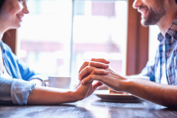 Cropped image of happy young couple holding hands, looking at each other and smiling while sitting at the cafe
