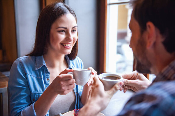 Happy young couple is drinking coffee, talking and smiling while sitting at the cafe