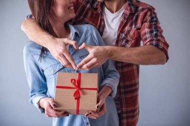 Mutlu genç gri arka plan üzerinde çift. Adam kalp gösterilirken kız bir hediye kutusu tutuyor, her ikisi de gülüyorsun