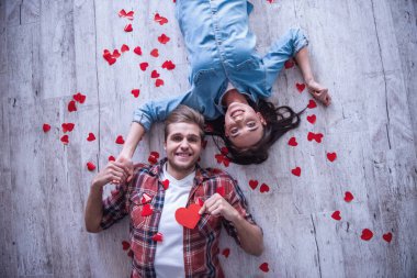 Üstten Görünüm mutlu genç çiftin kameraya bakarak ve ahşap yerde yatarken gülümseyerek. Adam kırmızı kağıt kalp tutuyor