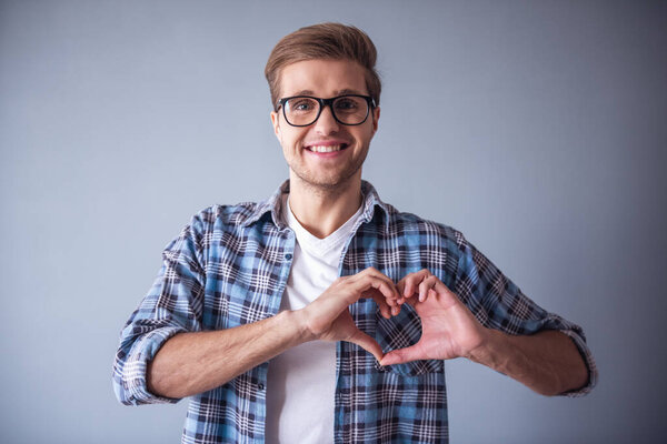 Красивый романтичный парень смотрит в камеру и улыбается, показывая сердце, на сером фоне
