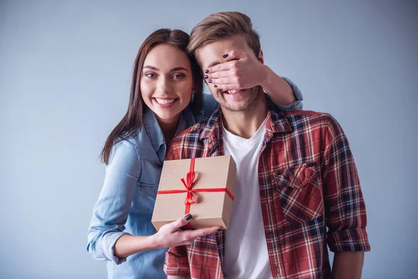 Beautiful girl is covering her boyfriend eyes and giving him a present, on gray background