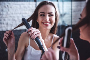 Güzel kız beyaz fanila Saç Düzleştirici kullanarak ve banyo aynada içine bakarken gülümseyen