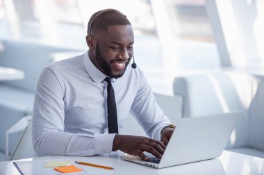 Yakışıklı Afro Amerikalı işadamı takım elbise ve kulaklık dizüstü bilgisayar Office ile çalışırken gülümsüyor