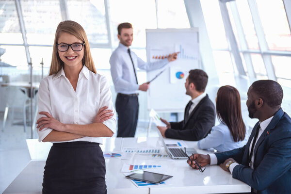 Деловая женщина в костюме смотрит на камеру и улыбается, деловая конференция на заднем плане
