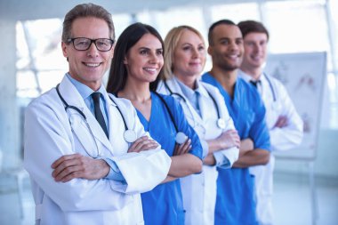 Tıp doktorları başarılı ekibi kameraya bakarak ve hastanede çapraz kolları olan dururken gülümseyen