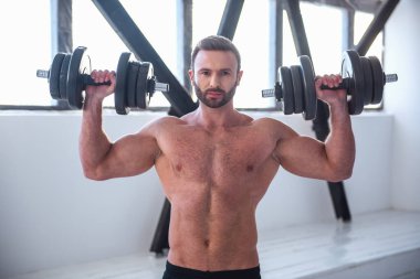 Çıplak gövde ile yakışıklı kaslı adam dumbbells ile dışarı çalışırken kameraya bakıyor