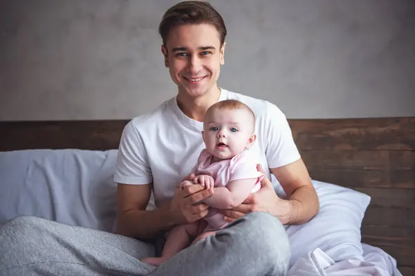 Handsome young dad is holding his cute baby in arms, looking at camera and smiling while sitting on bed