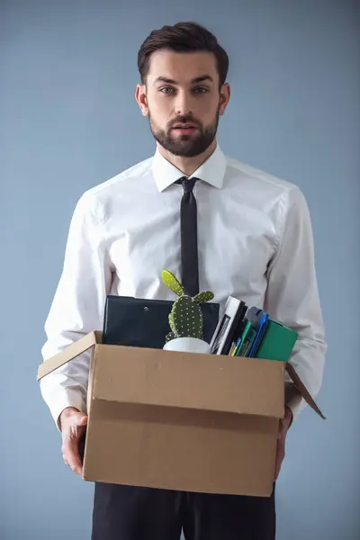 Getting fired. Handsome businessman in formal wear is holding a box with his stuff and looking at camera, on gray background