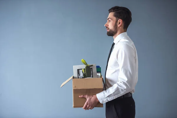 Getting fired. Side view of handsome businessman in formal wear holding a box with his stuff, on gray background