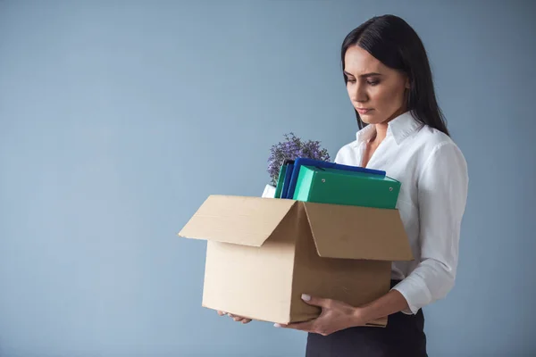 Getting fired. Beautiful young business woman in formal wear is holding a box with her stuff and looking sadly at it, on gray background