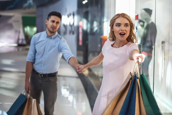Beautiful couple with shopping bags is doing shopping in the mall. Excited girl is pulling her tired boyfriend