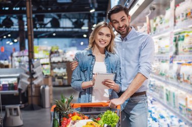 Güzel çift kameraya bakarak ve süpermarkette alışveriş yaparken gülümseyen