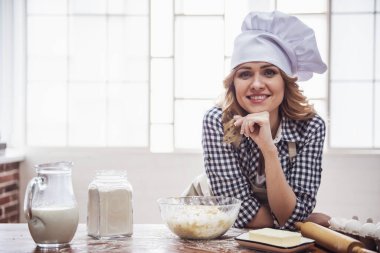 Önlük ve Şef şapkalı güzel genç kadın kameraya bakarak ve mutfakta hamur yoğurma sırasında gülümseyen
